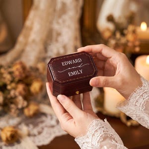May include: A dark burgundy, hexagonal ring box featuring a gold-toned latch and the white printed text "EDWARD and EMILY" on the lid, accented with a delicate gold border design around the top edge of the box.