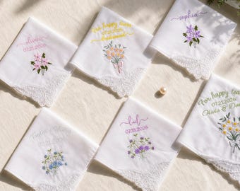 May include: Six white square handkerchiefs with scalloped lace edges, each featuring custom embroidery. Designs include floral bouquets in pink, yellow, purple, and blue, along with names and dates like 07.21.2026 and 07.18.2026, plus text reading For happy tears.