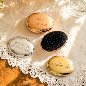 May include: Three oval hairbrushes with metallic finishes in rose gold, silver, and gold. One brush shows black bristles. Engravings include the names Sarah with date 06.15.2026, Will You Be My Bridesmaid?, and Love, Hannah in cursive script.