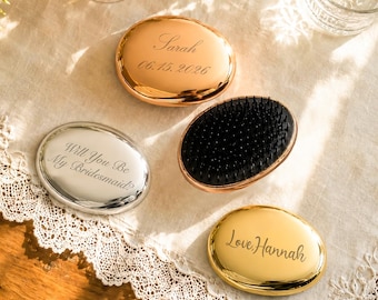 May include: Three oval hairbrushes with metallic finishes in rose gold, silver, and gold. One brush shows black bristles. Engravings include the names Sarah with date 06.15.2026, Will You Be My Bridesmaid?, and Love, Hannah in cursive script.