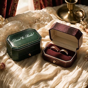May include: Two octagonal ring boxes, one dark green and one deep burgundy, featuring gold-tone latches. The green box lid is inscribed with the text "Daniel & Rachel" in white script. The open burgundy box contains two gold bands on a velvet insert.