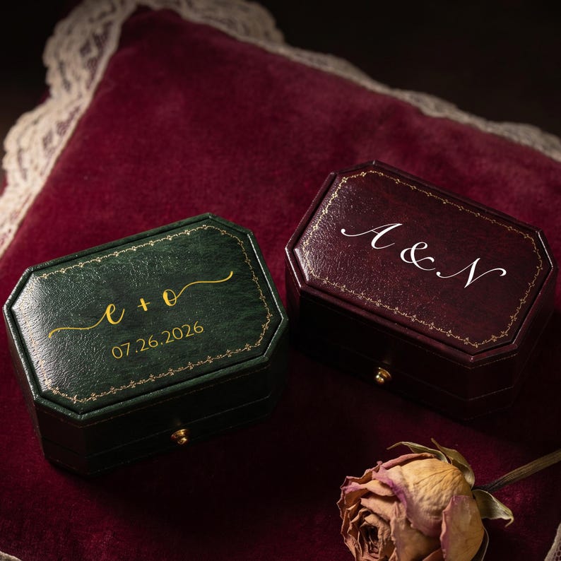 May include: Two octagonal ring boxes, one dark green and one deep burgundy, featuring gold-toned filigree borders. The green box displays the text e + o and 07.26.2026 in gold, while the burgundy box features the white script text A & N.