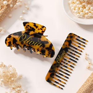 May include: A tortoiseshell-patterned hair claw clip and comb set. The clip is a rounded shape with teeth, and the comb has wide teeth. Both items have the name "Olivia" in gold script. The set is on a white surface with pearls.