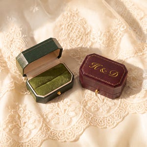May include: Two octagonal ring boxes, one dark green and one burgundy. The green box is open, revealing a green velvet interior. The burgundy box is closed and features gold lettering that reads H & D on the lid with a gold latch.