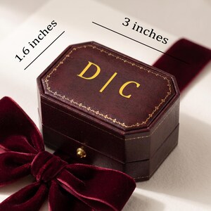May include: A dark burgundy, octagonal ring box featuring the gold-printed letters D|C on the lid, accented with a gold-coloured latch and a deep red velvet ribbon. The box measures 1.6 inches by 3 inches as indicated by the text.