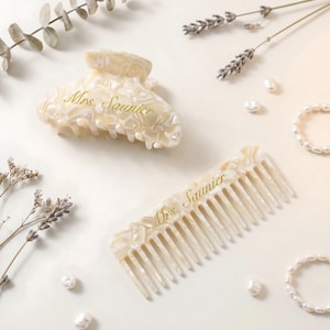 Pode incluir: Um conjunto de presilha e pente de cabelo branco sujo com o texto "Mrs. Saunier" em escrita dourada. A presilha tem formato arredondado e o pente tem dentes largos. O conjunto é rodeado por pérolas e raminhos de lavanda.