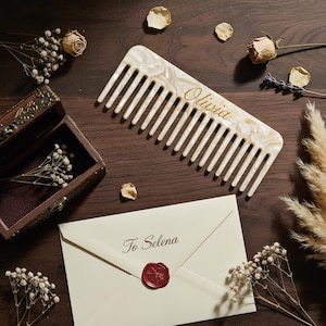 May include: A wide-tooth hair comb made of cream-colored marbled acrylic with the name Olivia printed in gold script. Below the comb is a cream-colored envelope sealed with red wax, featuring the text To Selena printed in dark brown script.