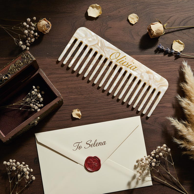 May include: A wide-tooth hair comb made of cream-colored marbled acrylic with the name Olivia printed in gold script. Below the comb is a cream-colored envelope sealed with red wax, featuring the text To Selena printed in dark brown script.
