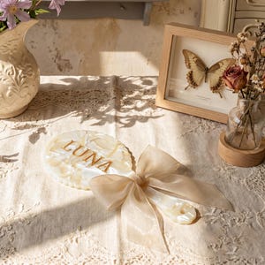 Op de afbeelding: Een handspiegel met een crèmekleurig, gemarmerd harsframe met de naam LUNA in gouden letters. Het handvat is versierd met een grote, doorschijnende, lichtbeige organza strik, wat zorgt voor een decoratief en functioneel accessoire.
