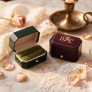 May include: Two rectangular ring boxes with octagonal edges. One box is dark green with a green velvet interior, and the other is dark maroon with a gold-coloured latch and the white printed letters D and C separated by a wheat stalk.