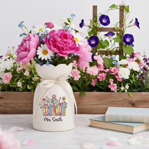 Könnte beinhalten: Eine cremefarbene Keramikvase mit Blumen- und Buchdesign, gebunden mit einem cremefarbenen Band, enthält einen Strauß aus rosa Pfingstrosen, Gänseblümchen und anderen Blumen. Die Vase ist mit dem Namen "Mrs. Smith" personalisiert. Ein Holzkasten mit Blumen und ein Spalier mit lila Blumen im Hintergrund.