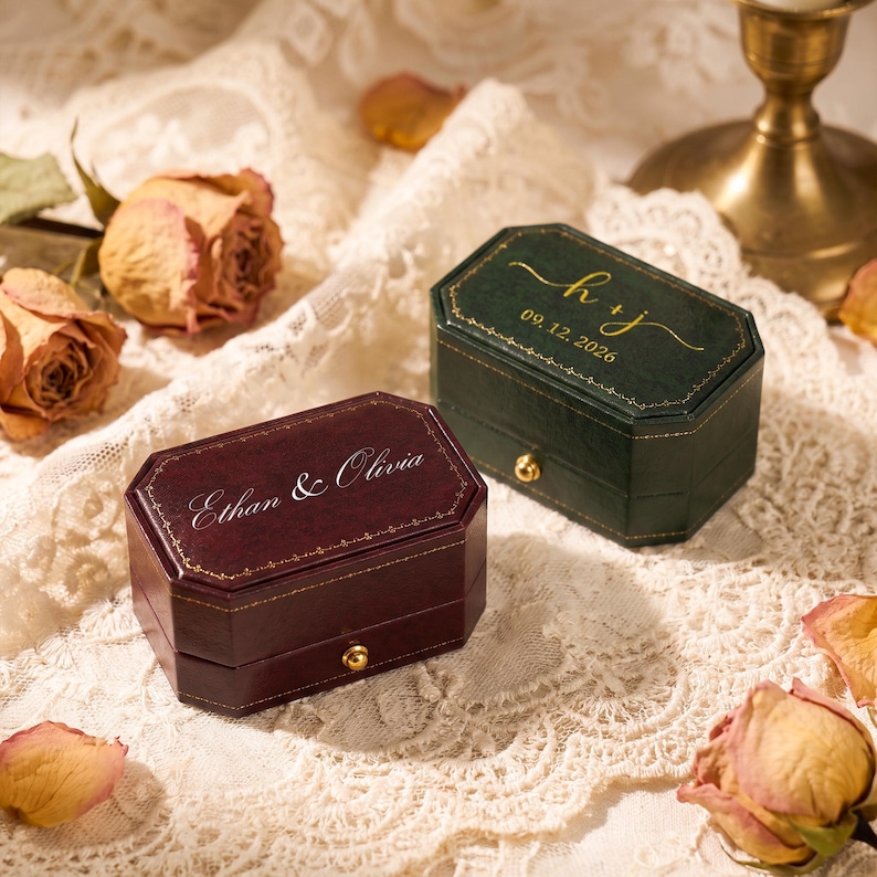 May include: A dark red rectangular ring box with the text Ethan & Olivia and a dark green rectangular ring box with the text h + j and 09.12.2026. Both boxes feature gold-coloured trim, a gold-coloured latch, and a smooth finish.