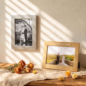 Puede incluir: Un marco de fotos vertical plateado grabado con Oliver + Sophia y 9.12.2026, y un marco de fotos horizontal dorado grabado con WILLIAM & CAMILA y EST. 2026. Ambos marcos contienen fotografías de bodas y se exhiben con rosas secas.
