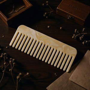 May include: A wide-tooth hair comb made of cream-colored marbled acetate with a smooth, polished finish. The comb features the name Olivia engraved in elegant gold cursive script on the center of the body, showcasing a classic rectangular shape.