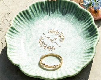 May include: A round, scalloped-edge ceramic trinket dish in a gradient of sage green and white. The centre features a gold-coloured floral wreath design with cursive lettering. A thin gold-toned metal ring with small clear stones rests inside.