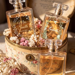 May include: Three rectangular glass perfume bottles with gold spray nozzles and clear caps, filled with amber liquid. The bottles feature custom white floral engravings and text including "Mrs. Smith 09.05.2026", "Isabella", and "Sept 16, 2026", nestled in a floral-patterned round box.