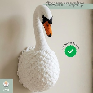 Op de afbeelding: Een witte gehaakte zwanentrofee met een lange nek en een rond lichaam. De snavel van de zwaan is oranje, met zwarte accenten. De tekst "Swan trophy" staat bovenaan de afbeelding en "Crochet Pdf Pattern" staat in een groene cirkel.