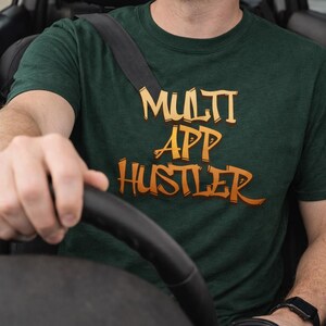 Może przedstawiać: Ciemnozielona koszulka z napisem "MULTI APP HUSTLER" w pomarańczowych i żółtych literach. Osoba ma zapięty pas bezpieczeństwa i trzyma kierownicę. Jednorazowy kubek do kawy stoi w uchwycie na kubek.