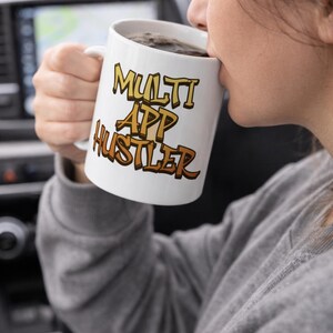 Może przedstawiać: Biały ceramiczny kubek z napisem "MULTI APP HUSTLER" w brązowej i żółtej czcionce. Kubek jest wypełniony ciemnym płynem, a osoba trzyma kubek.