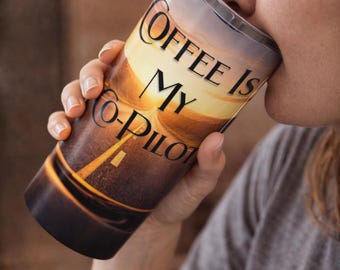 May include: A tall coffee tumbler with a sunset design and the text "Coffee Is My Co-Pilot." The tumbler is held by a person and is being brought to their mouth. The tumbler has a clear lid.