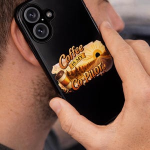 Może przedstawiać: Czarne etui na telefon z napisem "Coffee is my Co-Pilot" w stylizowanej czcionce. Projekt zawiera filiżankę kawy, wschód słońca i scenę leśną. Telefon jest trzymany przy uchu osoby.