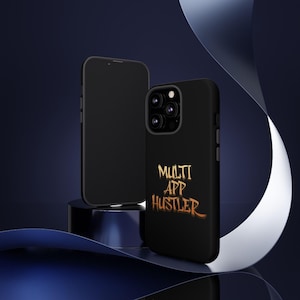 Puede incluir: Dos smartphones negros, uno con una funda protectora. La funda presenta el texto "MULTI APP HUSTLER" en una fuente dorada estilizada. Los teléfonos se muestran sobre una superficie oscura y reflectante, sobre un fondo azul oscuro.