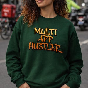 Może przedstawiać: Ciemnozielona bluza z napisem "MULTI APP HUSTLER" w pomarańczowych i żółtych literach. Bluza jest połączona z niebieskimi dżinsami. Osoba stoi na ulicy.