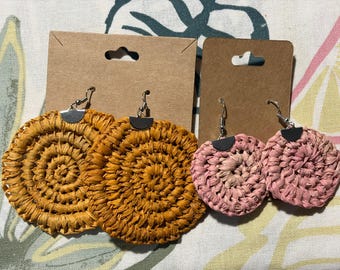May include: Two pairs of handmade raffia earrings. One pair is golden yellow, the other is pink. Both have a circular, spiral design and silver-tone hooks. They are displayed on brown earring cards.
