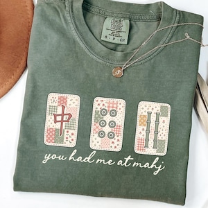Puede incluir: Una camiseta de manga corta color verde oliva con un gráfico de tres fichas de mahjong con patrones florales de patchwork. Las fichas muestran un carácter chino rojo, seis círculos y tallos de bambú verdes. El texto dice, you had me at mahj.