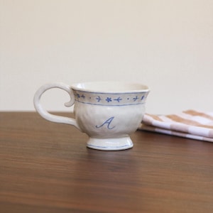 May include: A white ceramic teacup with a blue floral design around the rim and a blue letter "A" on the front. The teacup has a curved handle and sits on a dark wooden surface. A folded cloth is in the background.