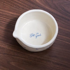 May include: A small, round, off-white ceramic bowl with a small pouring spout on the left side. The interior of the bowl features the blue handwritten text "Life is good" centered at the bottom, with small blue floral accents on the exterior.