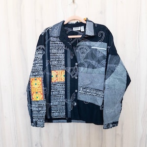 May include: A black long-sleeve button-down shirt featuring a patchwork design with various gray and white geometric patterns, floral line art, and a single rectangular panel of bright yellow and orange fabric on the left sleeve and front.