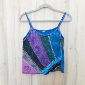 May include: A sleeveless tank top featuring a patchwork design of diagonal fabric panels in shades of blue, purple, black, and green. The garment is decorated with printed paisley and Om symbols, accented by visible white decorative stitching.