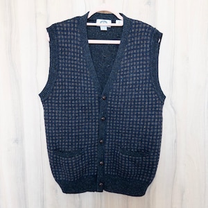 May include: A navy blue and brown checkered pattern sweater vest with a V-neck, two front pockets, and button closure. The vest is displayed on a pink hanger against a white background.