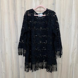 Może przedstawiać: Czarny top z koronki z frędzlami. Top wykonany jest z delikatnej, ażurowej koronki i ma długie rękawy i okrągły dekolt. Dolny brzeg wykończony jest frędzlami.