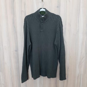 May include: Dark green ribbed knit sweater with a mock neck and three-button closure. The long-sleeved sweater is hanging on a white hanger against a light wood-grain background. The sweater has a green collar.