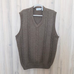 May include: A brown cable knit sweater vest with a V-neck. The sleeveless vest hangs on a grey hanger. The sweater has a ribbed hem and cable knit detailing down the front.