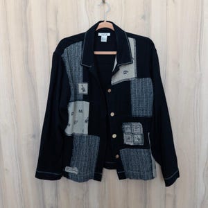 May include: A long-sleeved black button-down jacket featuring a patchwork design. The garment incorporates contrasting panels of textured grey fabric and light grey sections adorned with small, dark animal illustrations, finished with visible stitching.