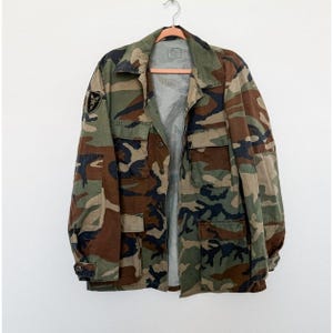 Könnte beinhalten: Tarnmusterjacke mit mehreren Taschen und Knopfleiste. Die Jacke hat ein grünes, braunes und schwarzes Tarnmuster. Ein Aufnäher ist auf dem linken Ärmel sichtbar. Die Jacke hängt an einem Holzkleiderbügel.