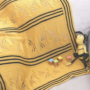 May include: A yellow knitted baby blanket with black stripes and a bee pattern. The blanket is on a white couch with a bee plush toy and colorful blocks.