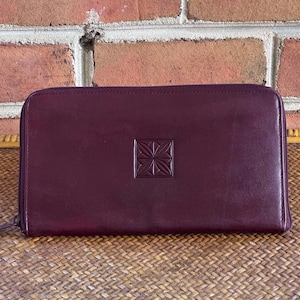 May include: A rectangular, deep burgundy leather wallet with a wrap-around zipper closure. The center of the wallet features a square embossed design consisting of eight symmetrical triangular segments arranged in a geometric star-like pattern.
