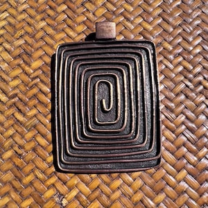 May include: A rectangular bronze-toned pendant featuring a raised, continuous spiral design that winds inward toward the center. The metal surface has a dark, textured finish with lighter highlights along the edges of the raised spiral pattern.