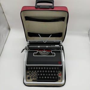 May include: A black and silver Olivetti Lettera 32 typewriter housed in a red carrying case with a black handle. The typewriter features a QWERTY keyboard layout, a metal carriage, and a visible ribbon spool cover. The case interior is beige.