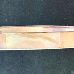 May include: A circular bangle bracelet featuring a polished metallic band inlaid with a smooth, iridescent panel of light peach and cream-colored shell material that displays a subtle, swirling pearlescent texture across its surface.