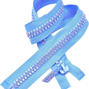 Puede incluir: Una cremallera azul claro con una fila de brillantes pedrería a cada lado. El tirador de la cremallera también es azul claro. La cremallera se muestra en forma curva sobre un fondo blanco.