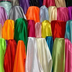 May include: A collection of numerous fabric bolts arranged in a dense display. The material features a smooth, reflective satin finish in a wide spectrum of vibrant colors including red, orange, yellow, green, blue, purple, pink, white, and silver.