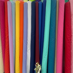 May include: A display of fabric rolls in a variety of colours, such as pink, red, orange, yellow, and blue. The rolls are neatly arranged, showcasing a wide array of textiles. A vase with flowers is in front of the fabric rolls.