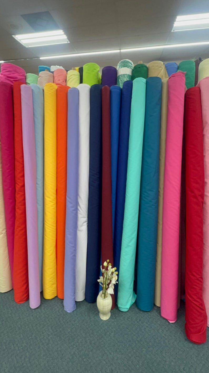 May include: A display of fabric rolls in a variety of colours, such as pink, red, orange, yellow, and blue. The rolls are neatly arranged, showcasing a wide array of textiles. A vase with flowers is in front of the fabric rolls.
