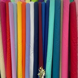 May include: A display of fabric rolls in a variety of colours, such as pink, red, orange, blue, and green. The rolls are standing vertically, highlighting the different colours and textures. A vase of flowers is placed in front of the rolls.
