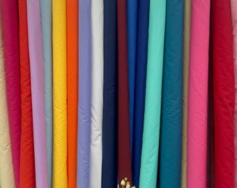 May include: A display of fabric rolls in a variety of colours, such as pink, red, orange, blue, and green. The rolls are standing vertically, highlighting the different colours and textures. A vase of flowers is placed in front of the rolls.